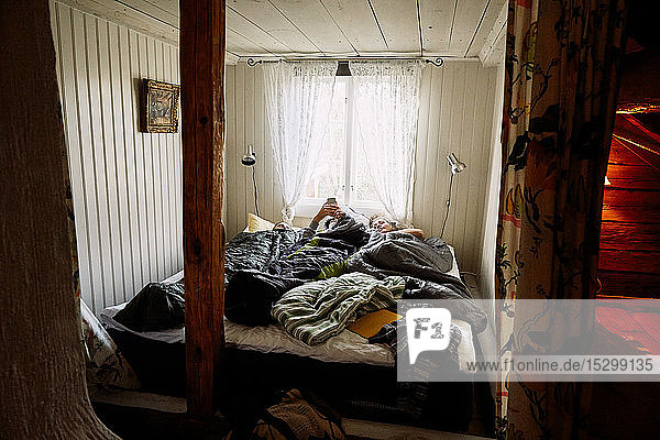 Junger Mann benutzt Smartphone  während er mit Freundinnen im Bett in einer Hütte liegt