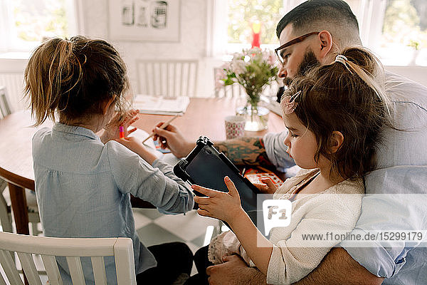 Mädchen benutzt digitales Tablett  während der Vater der Tochter bei den Hausaufgaben am Esstisch hilft