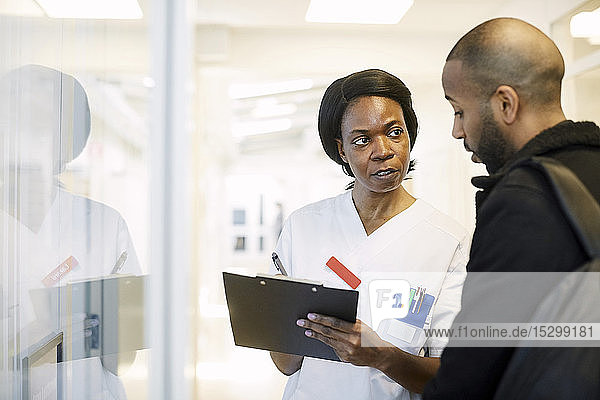 Ärztin diskutiert mit männlichem Patienten über Klemmbrett im beleuchteten Flur des Krankenhauses
