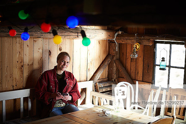 Portrait of smiling young woman using smart phone while sitting at table in log cabin