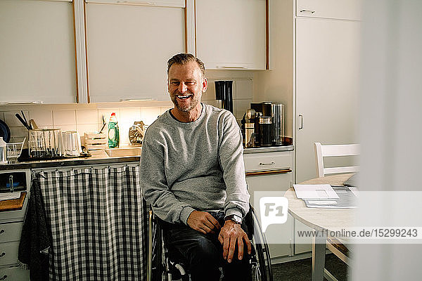 Fröhlicher reifer Mann sitzt zu Hause in der Küche auf dem Rollstuhl