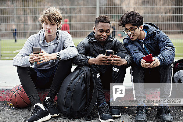 Social media addicted friends using mobile phones while sitting on sidewalk after basketball practice in city