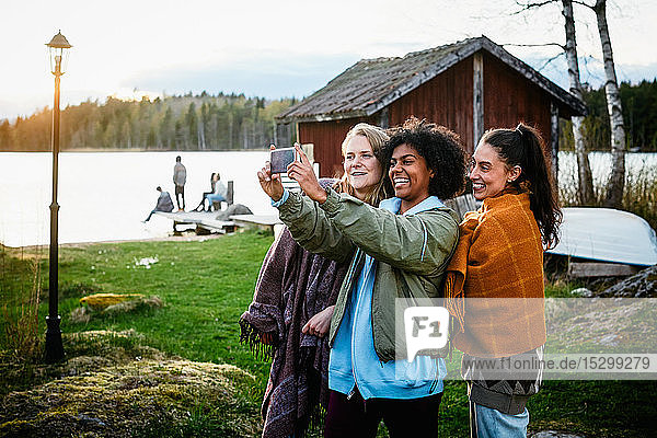 Glückliche Freundinnen  die sich mit einem Smartphone selbstständig machen  während sie bei Sonnenuntergang auf einem Grasfeld am See stehen