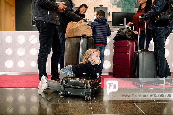 Boredom girl looking away while leaning on suitcase with family standing at reception in hotel