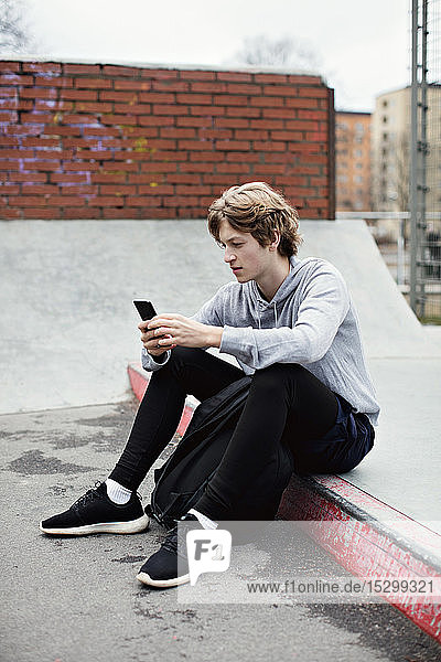 Full length of teenage boy using smart phone while sitting on street in city