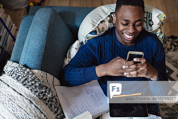 Schrägansicht eines sozialmediensüchtigen Teenagers  der ein Smartphone benutzt  während er auf dem Sofa mit Büchern im Wohnzimmer liegt