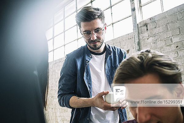 Selbstbewusster junger männlicher Computerprogrammierer hält Kaffeetasse in der Hand und steht einem Mitarbeiter im Kreativbüro zur Seite