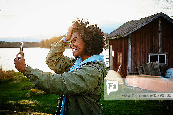 Glückliche junge Frau  die bei Sonnenuntergang am See steht und sich mit einem Smartphone selbstständig macht