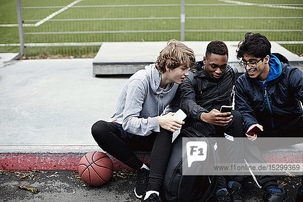 Männliche Freunde benutzen Mobiltelefone  während sie nach dem Basketballtraining im Winter auf dem Bürgersteig sitzen