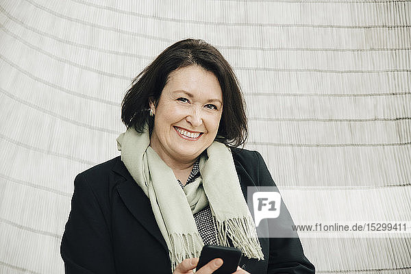 Porträt einer lächelnden  reifen Geschäftsfrau mit einem Smartphone in der Hand  die im Büro an der Wand steht