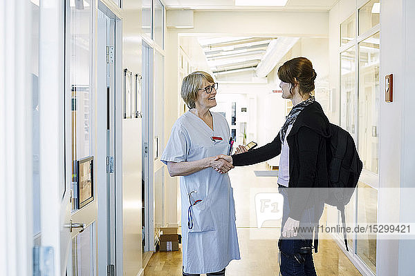 Smiling female practitioner shaking hands with patient during routine visit in hospital corridor