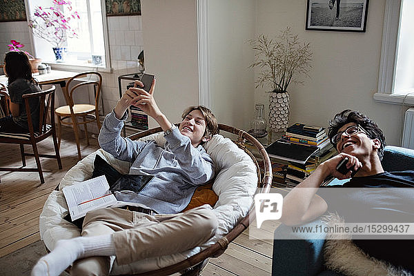Smiling teenage boy taking selfie while studying with friend in living room at home