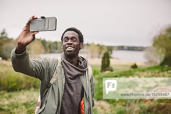 Smiling young man taking selfie with smart phone while standing on landscape against sky