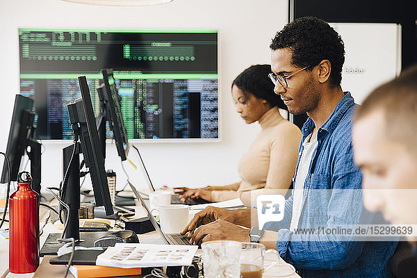 Seitenansicht von männlichen und weiblichen Computerhackern  die Laptops am Schreibtisch benutzen  während sie im Büro sitzen