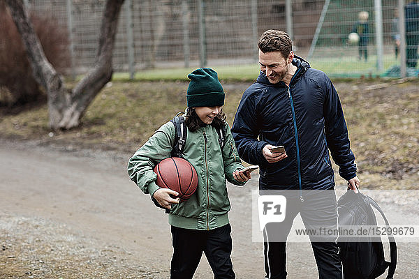 Happy father and son using mobile phones while walking on street after basketball practice in winter