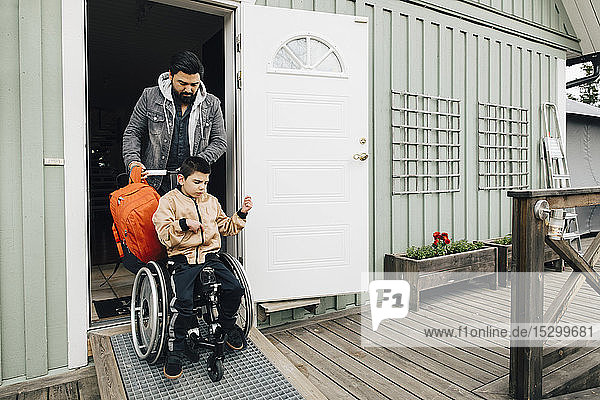 Vater mit Rucksack schiebendem autistischen Sohn im Rollstuhl vor der Tür