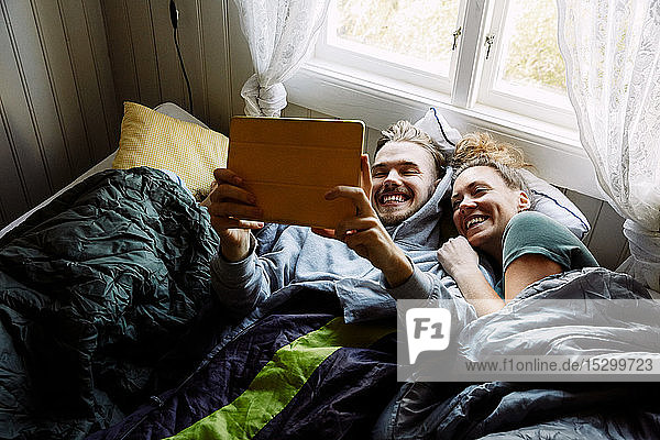 Hochwinkelansicht von lächelnden Freunden  die sich über ein digitales Tablet einen Film ansehen  während sie in einem Ferienhaus auf dem Bett liegen