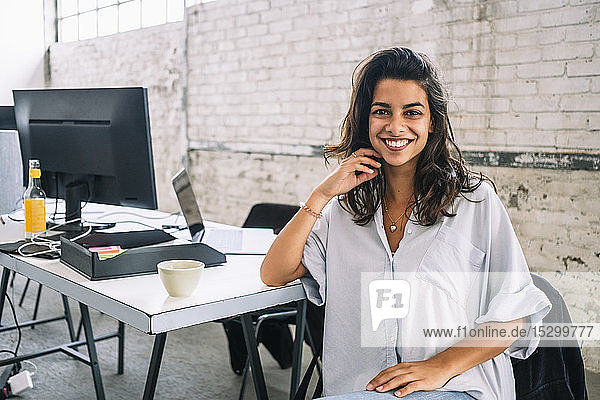 Portrait of smiling young female computer hacker sitting at desk in creative office
