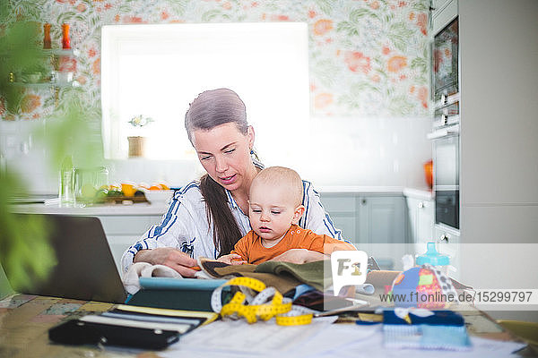 Modedesignerin kümmert sich um ihre Tochter  während sie zu Hause am Tisch in der Küche arbeitet