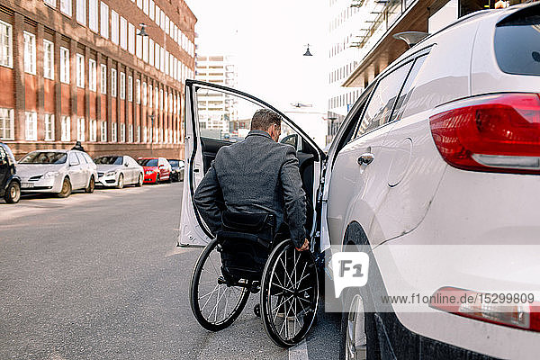Rear view of mature man boarding white car while sitting on wheelchair in city
