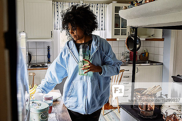 Junge Frau mit lockigem Haar hält Trinkgläser  während sie zu Hause in der Küche steht