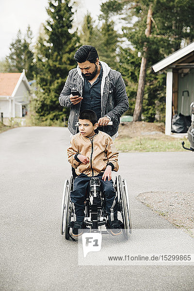 Vater benutzt Smartphone  während er seinen autistischen Sohn im Rollstuhl schiebt