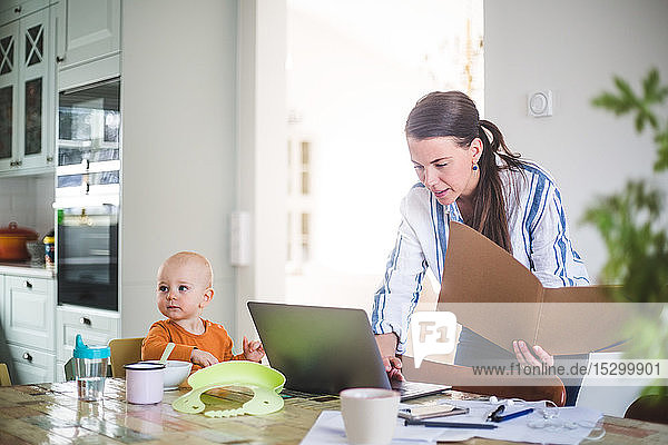 Weibliche Berufstätige hält Akte  während sie den Laptop ihrer Tochter auf dem Esstisch im Heimbüro benutzt