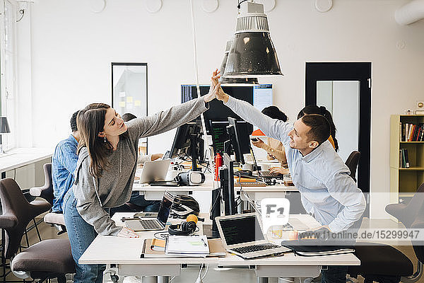 Happy male and female computer programmers doing high-five over computer at desk in creative office
