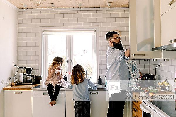 Girl looking at working sister while father doing chores in kitchen