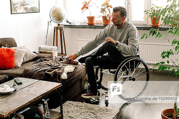 Full length of disabled mature man wearing shoe while sitting on wheelchair in living room at home