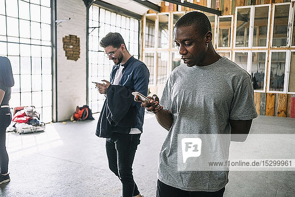 Selbstbewusster junger Geschäftsmann benutzt ein Smartphone  während er am kreativen Arbeitsplatz an einem Kollegen vorbeigeht