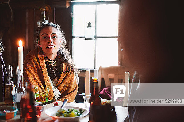 Junge Frau mit Decke schaut weg  während sie am Tisch gegen das Fenster im Blockhaus sitzt