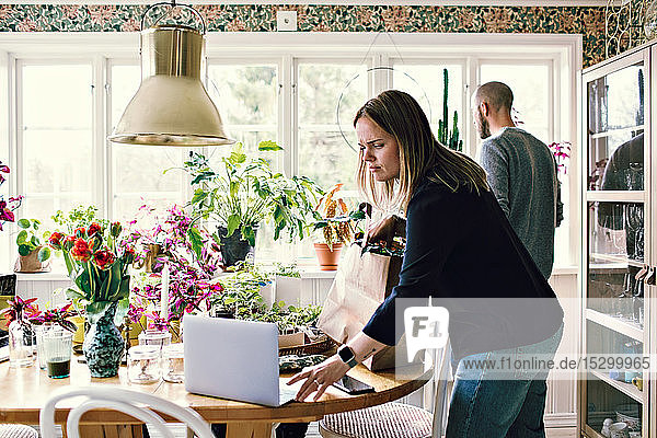 Woman using laptop at table while man gardening in room