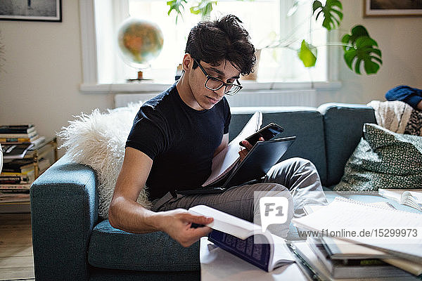Serious young man doing homework while sitting on sofa with mobile phone and laptop at home