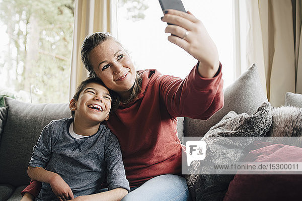 Mother taking selfie with happy autistic son while sitting on sofa at home