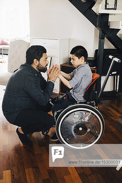 Side view of father holding autistic son's hands sitting on wheelchair at home