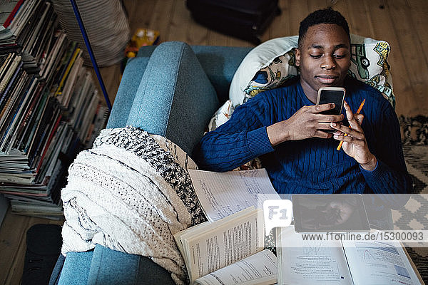 Schrägansicht eines Teenagers  der ein Mobiltelefon benutzt  während er zu Hause mit Büchern auf dem Sofa liegt
