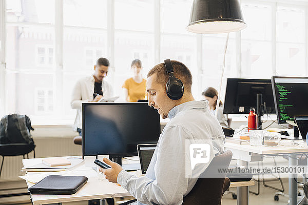 Seitenansicht eines männlichen Ingenieurs  der ein Mobiltelefon benutzt  während seine Kollegen im Hintergrund an einem kreativen Arbeitsplatz arbeiten