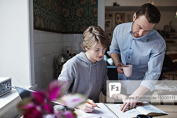 Vater hält Kaffeetasse in der Hand  während er dem Sohn zu Hause beim Lernen hilft