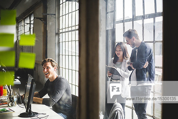 Fröhlicher Geschäftsmann sitzt am Schreibtisch  während IT-Fachleute im Hintergrund durch Glas gesehen arbeiten