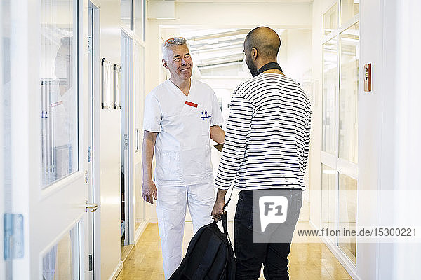 Smiling healthcare worker looking at male patient during visit in hospital corridor