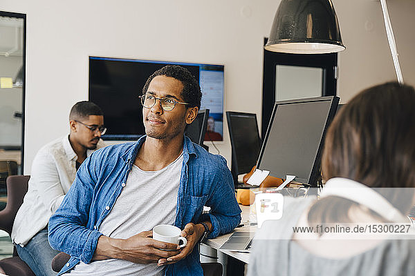 Nachdenklicher männlicher Computerprogrammierer schaut weg  während er neben Kollegen am Schreibtisch im Büro sitzt