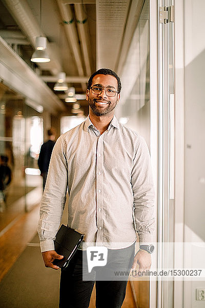 Porträt eines lächelnden  mittelgroßen Geschäftsmannes  der ein digitales Tablet im Bürokorridor hält