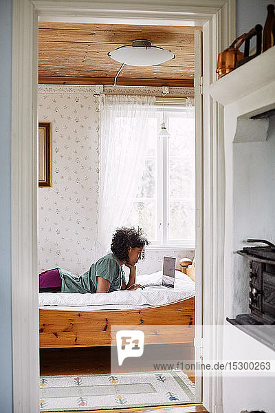 Seitenansicht einer jungen Frau  die einen Laptop benutzt  während sie auf dem Bett liegt  gesehen durch eine Türöffnung zu Hause