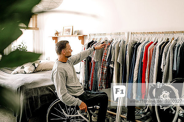 Älterer Mann sitzt auf Rollstuhl  während er zu Hause Kleider vom Ständer auswählt