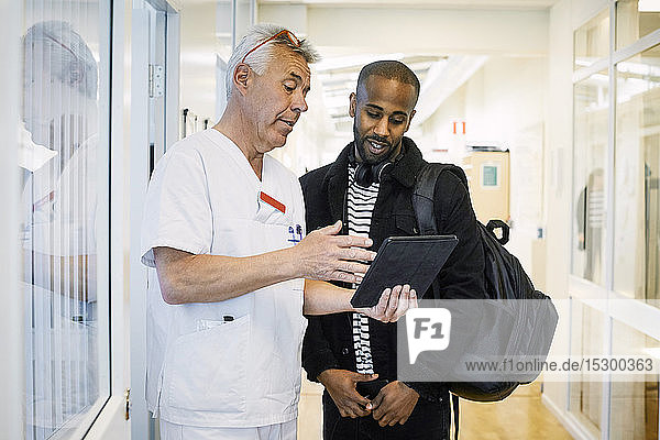 Senior doctor discussing with young man over digital tablet during routine check up in corridor