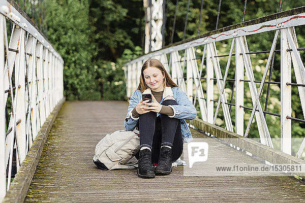 Teenager-Mädchen sitzt auf einer Brücke und benutzt ein Smartphone