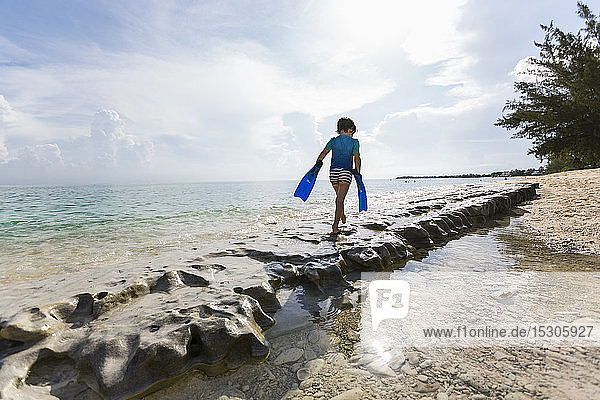 5-jähriger Junge  der mit Schwimmflossen auf Felsen am Ufer läuft