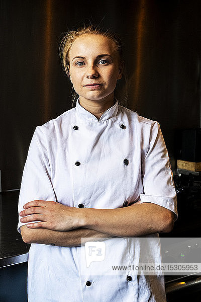 Woman wearing chef's jacket standing indoors with her arms folded  looking at camera.