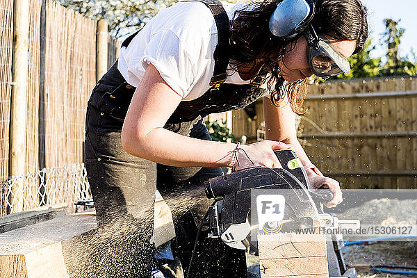 Woman wearing protective goggles and ear protectors holding circular saw  cutting piece of wood on building side.